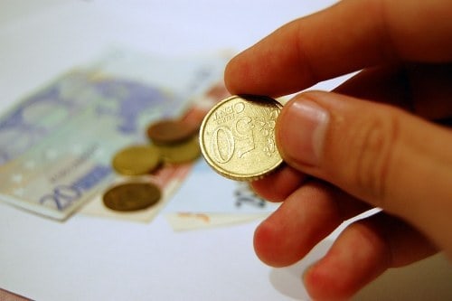Close-up of hand holding a €1 coin with euro banknotes and coins in the background.