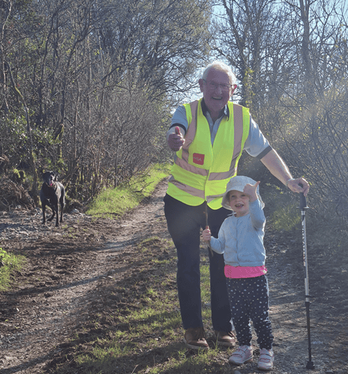 Elderly man and girl walking outdoors with dog on nature trail in Ireland.